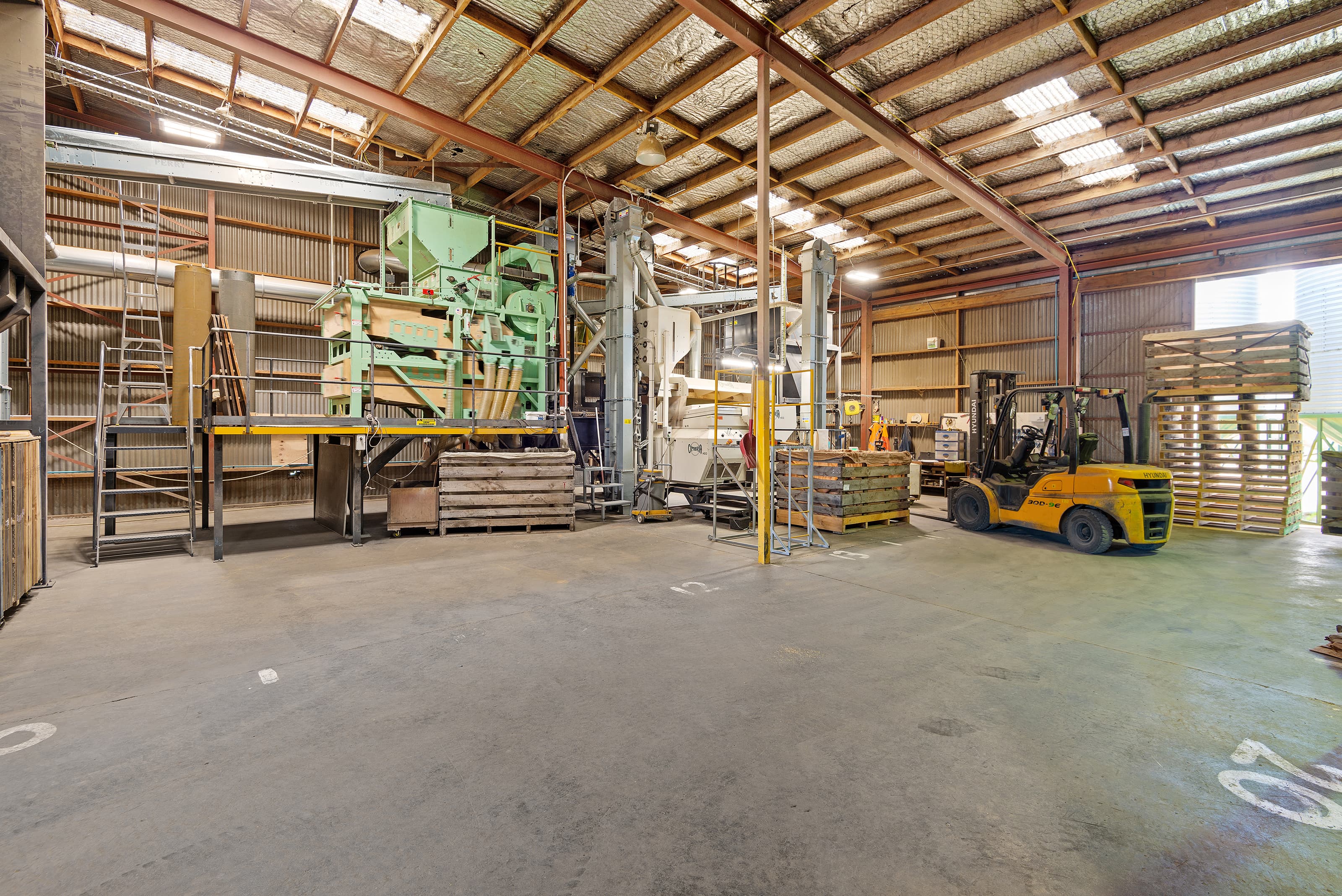 Industrial warehouse interior with large processing machinery, a yellow forklift, and stacked wooden pallets.