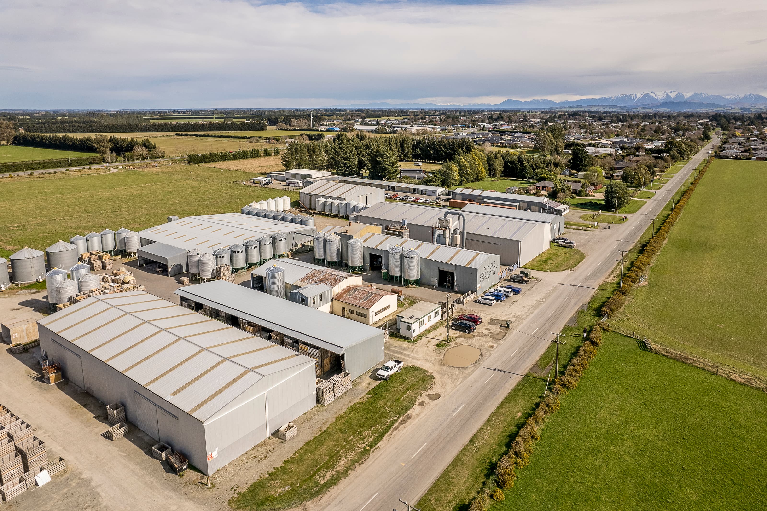 Aerial view of a seed cleaning facility with silver silos, warehouses, and distant mountains.