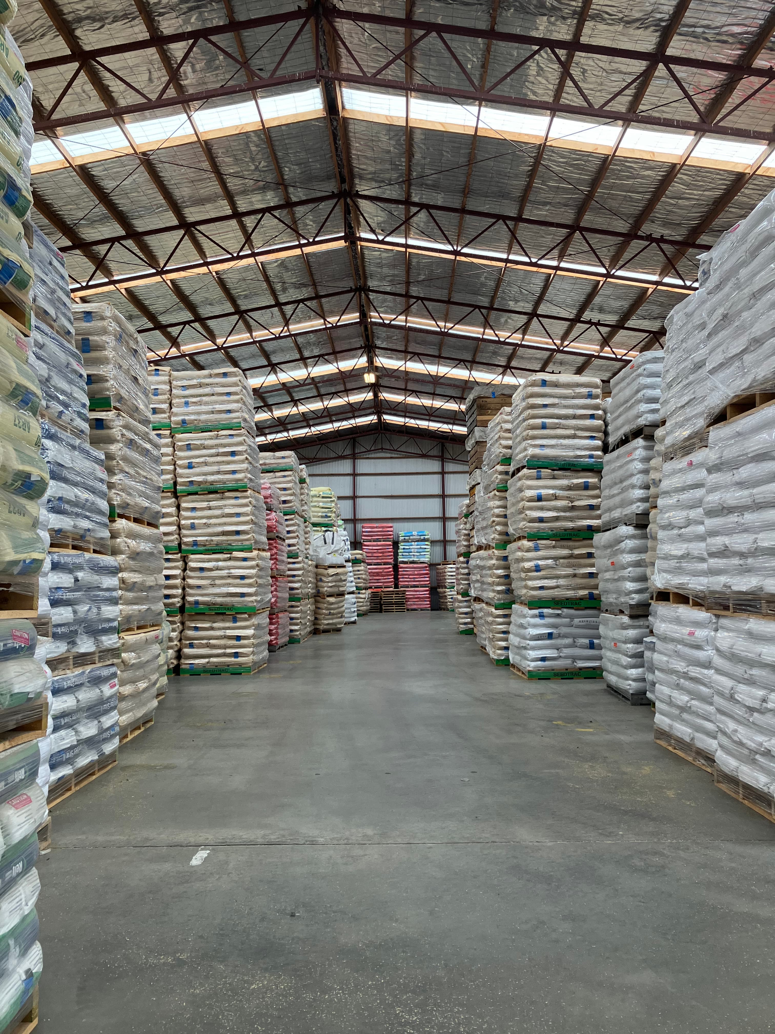 Pallets of shrink-wrapped bags stacked high in a large warehouse with a metal truss ceiling.