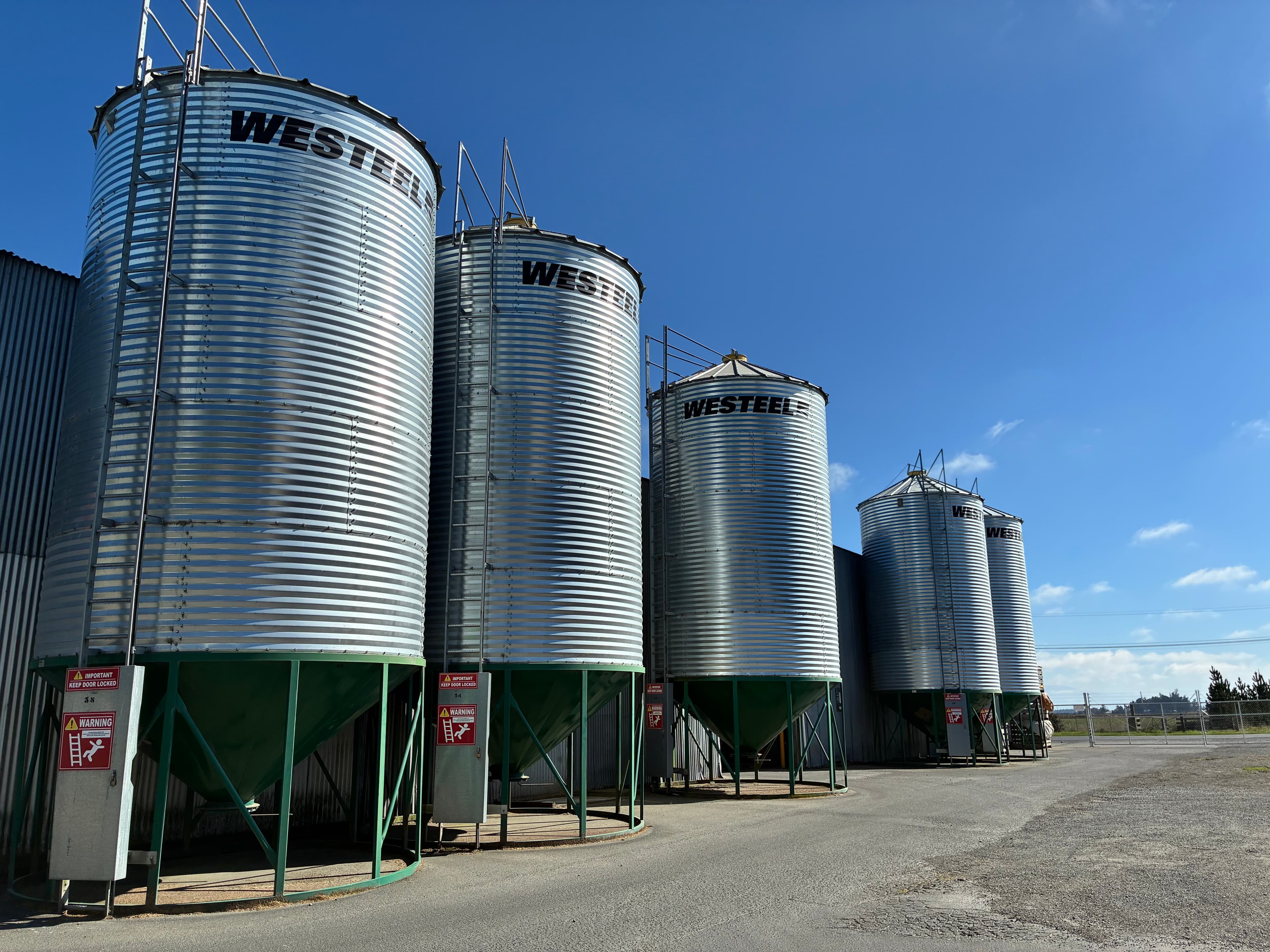 Large corrugated metal Westeel grain silos with green conical bases under a blue sky.